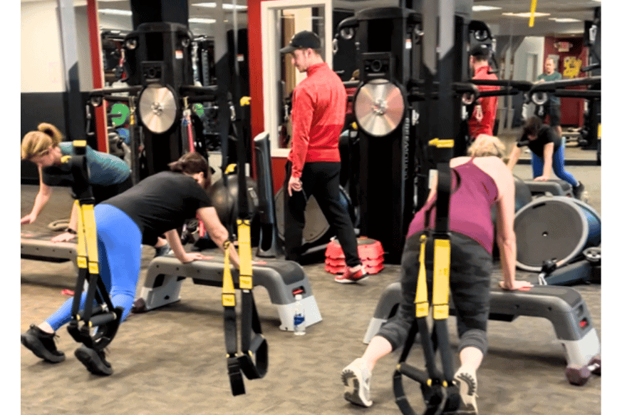 small group training class with 3 women & a personal trainer showing various equipment used like step platform & trx bands