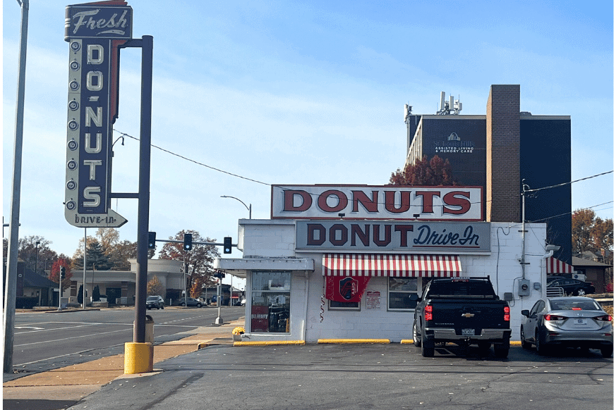 see the exterior of Donut Drive In, a tiny donut shop with large light up signs and a red and white striped banner, parking lot