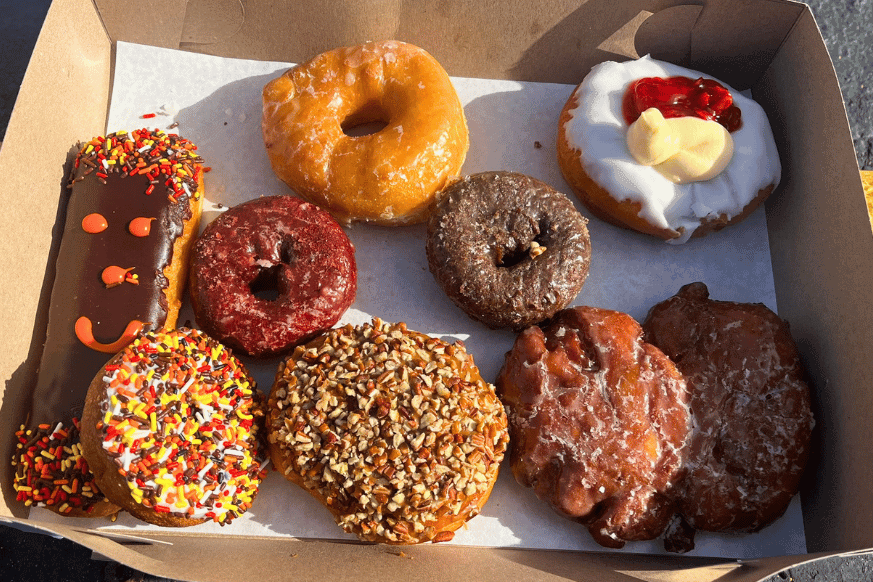 box of donuts with a variety of toppings  from Donut Drive In includes a chocolate long john with sprinkles, an apple fritter, a caramel pecan cinnamon roll, chocolate glazed, red velvet glazed donuts.