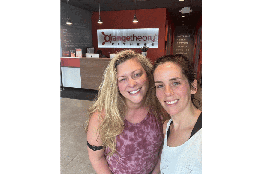 two women smiling at the orangetheory fitness center in ladue, MO.  women on the left is wearing a heart rate monitor which helps track your performance statistics during class.