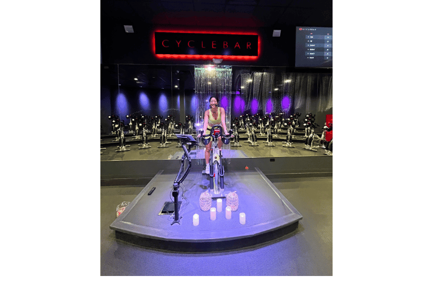 a smiling cyclebar instructor, on top of her stationary bike on stage in the front of the room at the creve coeur location of cyclebar.