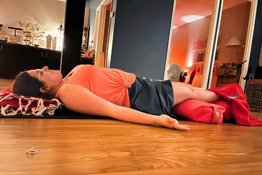 A women with brown hair and glasses looks peaceful and restful while practicing Yoga Nidra at a yoga studio.  She is lying down on the hardwood floor with pillows under her knees and blankets on her feet.  Her head is propped up with a blanket to use as a pillow. 