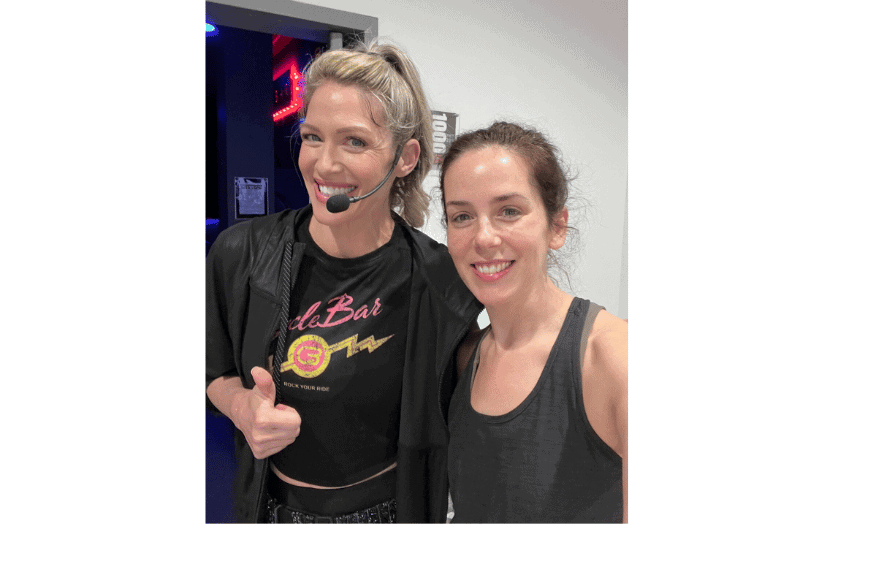 2 women, one instructor with headset microphone and one class attendee, smiling & giving a thumbs up before entering a cyclebar class