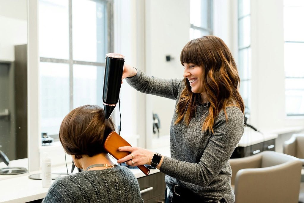 inside of a beauty salon, a women with short brown hair is seated in a swival chair facing a mirror.  She is having her hair blown dry by a female hairdresser with long brown wavy hair.  