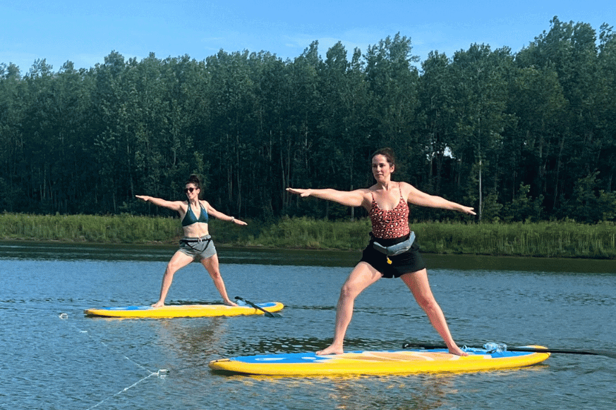 Natalie Kalmar and friend standing on large yellow and blue paddleboard in warrier 2 yoga pose in the middle of the lake at River's Edge Park.  This fitness event is hosted by SUP St. Louis.