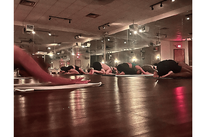 inside Sumits Hot Yoga studio with mirrored walls & hardwood floors.  Several yogis are resting on their mat in Child's Pose.   