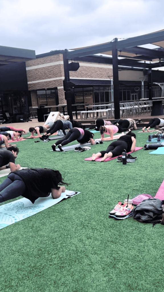 Participants attending a Mat Pilates class by Club Fitness, hold a plank pose on their yoga mats at The District In Chesterfield, MO.  Brightly colored yoga mats are spread out on the astroturf in the center of The District. 