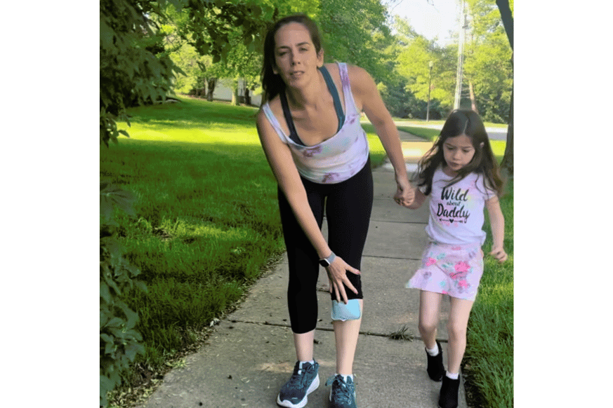 A mother women walking on the sidewalk with her young daughter, wincing in pain and holding her knee. 