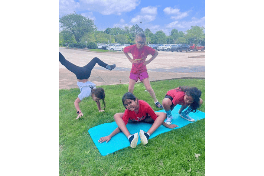 4 young girls, roughly 8 years old, each doing a different yoga pose outside of an elementary school.  they are on the grassy area near a school parking lot with a turquoise yoga mat.