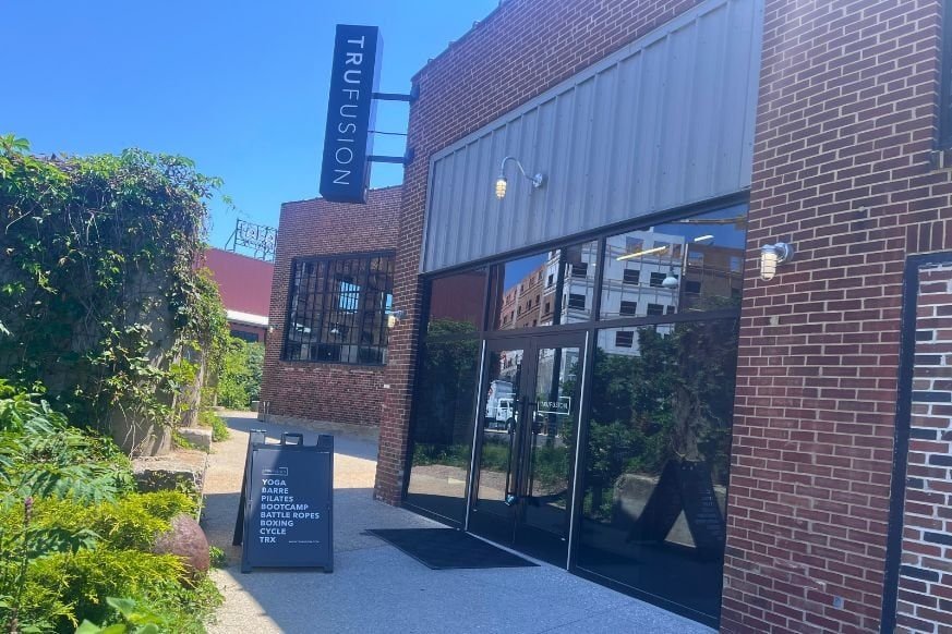 Brick building with large glass doors and windows feature the TruFusion logo.  Outside lush greenery marks the sidewalk and a triangle sign showcases TruFusions class offerings.