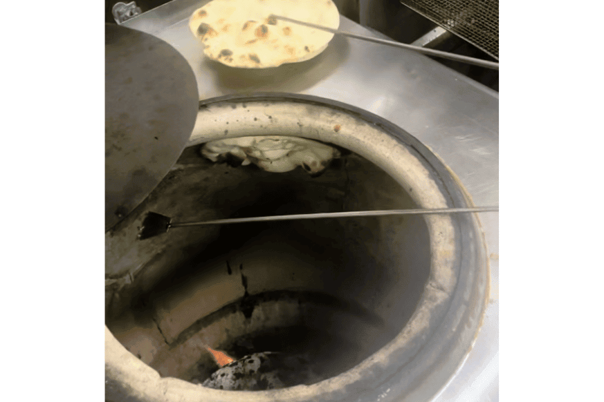 clay cylindrical cooking device shown with flame and charcoals at the bottom. long poker took is being used to remove naan from the walls.