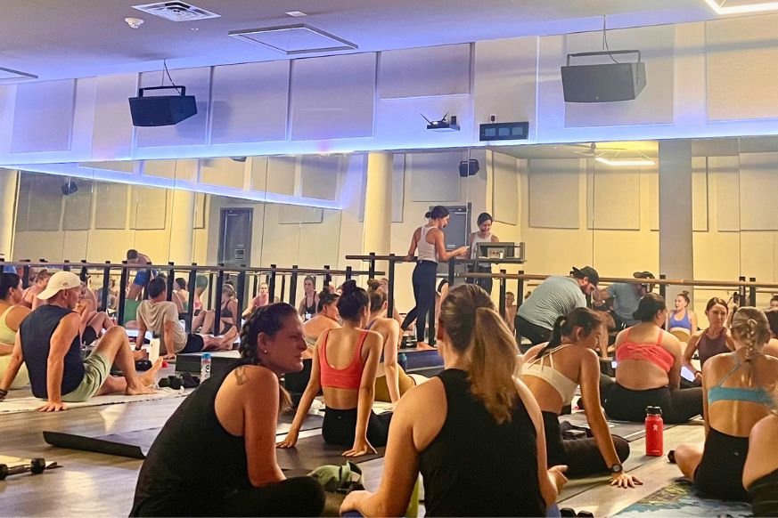 very crowded exercise room with mirrored walls and a ballet barre.  many men & women in athletic wear seated on black mats waiting for class to start.  instructor is standing at the front of the room, adjusting the music to start class. 
