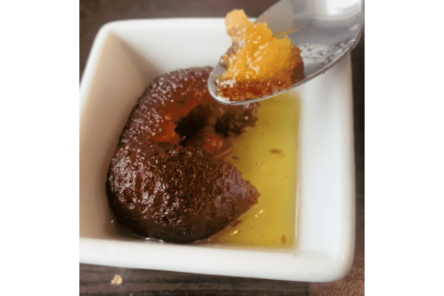 white porcelain dish with a dark brown, oval shaped dumpling thats been cut open with a spoon.  spoon contains a small amount of the dessert that is golden in color.  the dumpling is resting in a sugary honey colored liquid.