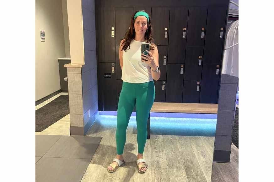 FitnessFoodieSTL, aka Natalie Kalmar, standing in the TruFusion St Louis locker room taking a mirror selfie.  She is wearing green yoga pants with matching green headband, white tank and sandles.  floor lit locker rooms with wooden bench.  