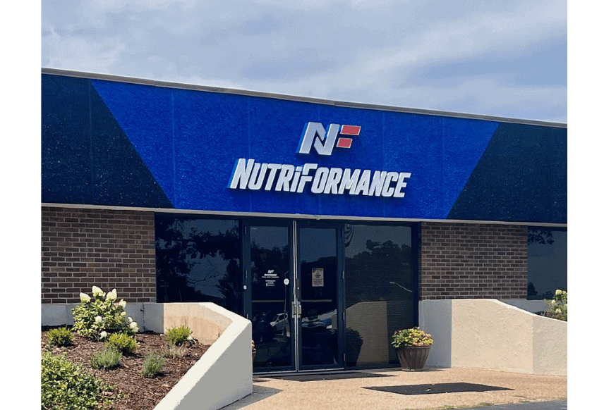 exterior entrance to NutriFormance in Creve Coeur, a suburb in St. Louis, MO.  blue background with white & red logo.  flowers and plants frame the exterior doors. 