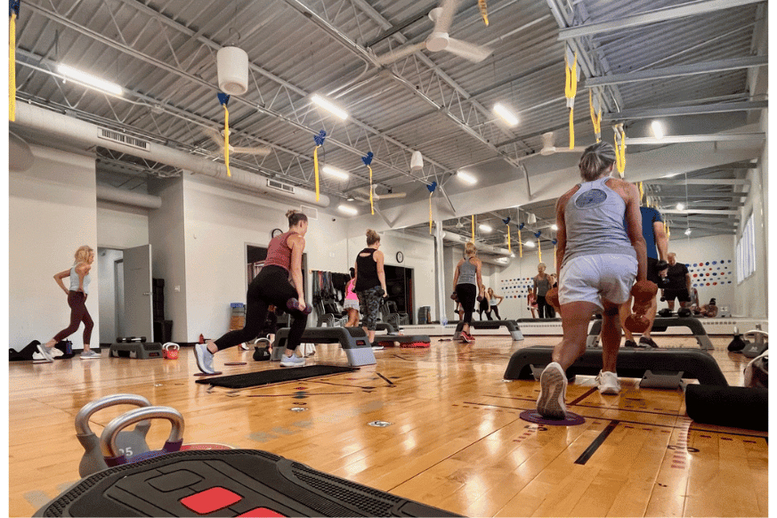 Busy group fitness class shows mixed ages lunging with weights alongside other eqipment like steppers, yoga mats and kettleballs. 