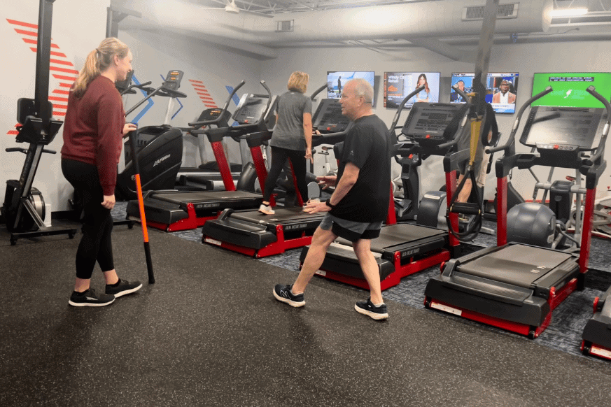 cardio equipment like treadmills, stair climbers, and ellipticals with televisions in the background.  in the foreground and elderly man working out with a young personal trainer. 