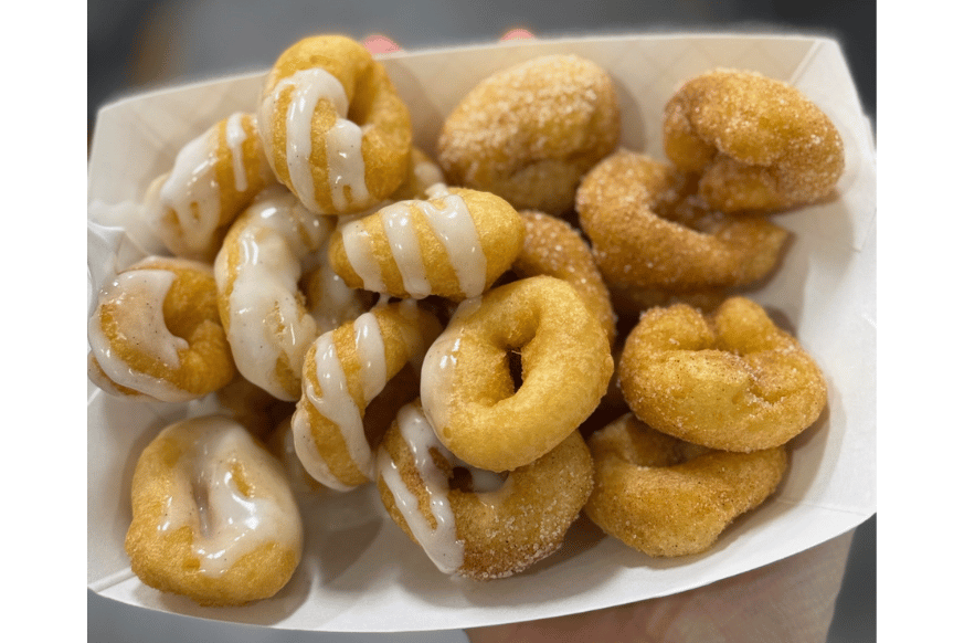 small round donuts piled high in a white container.  half are drizzled with a white sugary glaze and the other half are topped with cinnamon and sugar