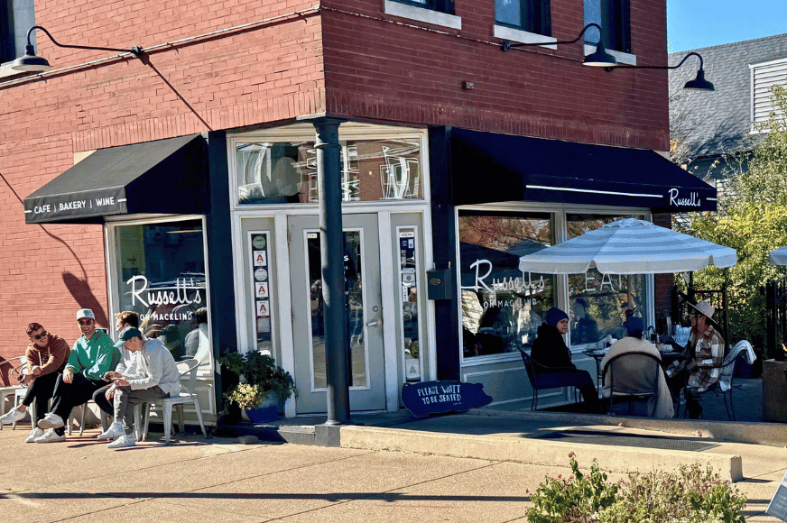 The sunny street corner of Russell's on Mackland. 2 Black awnings, outside tables with umbrellas and chairs for guests waiting for an indoor table.  