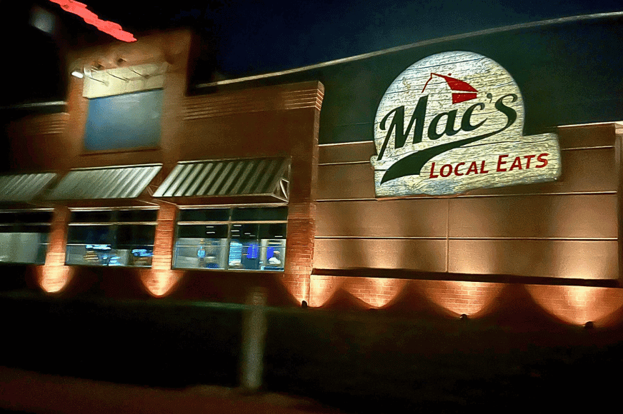 the outside of Mac's local eats at night with dramatic uplighting showcasing their logo  and green and white striped awnings on the windows.  