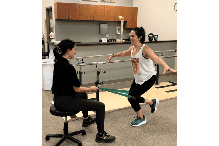 female patient with a knee injury is balancing on one leg while physical therapist uses resistance band to engage the patients bad knee