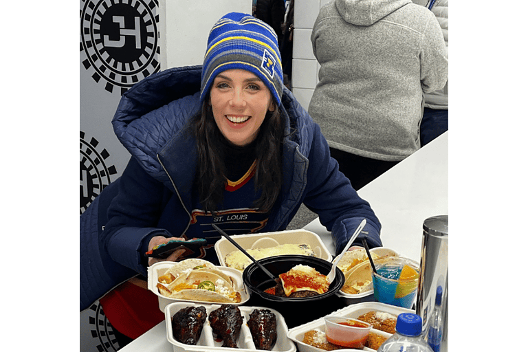 natalie kalmar poses with a large platter of enterprise center concessions from bret hull's junction house. natalie is dressed in a blue coat and blues winter hat. food shown includes chicken drumsticks, big-ass meatball and spinach artichoke bread