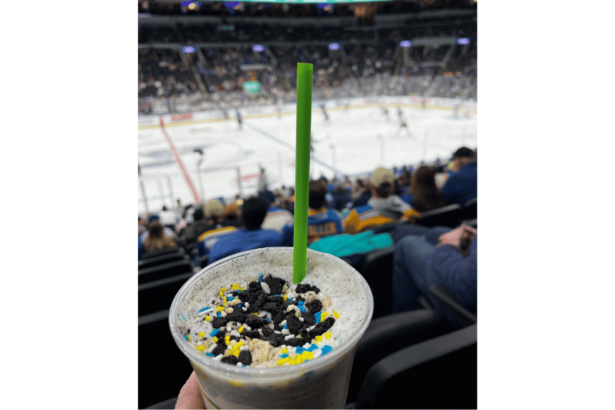 St. Louis Blues game in the background and Shake Shack's MVP shake shown, cookies and cream shake topped with cookie crumbs and yellow, white and blue sprinkles. green straw.