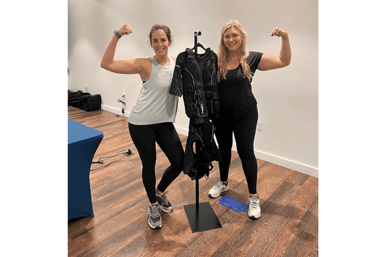 two women hold a bicep curl pose near an EMS body suit