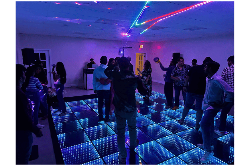 people dancing in dimly lit large room with multi colored lights.  dj in front of room.  