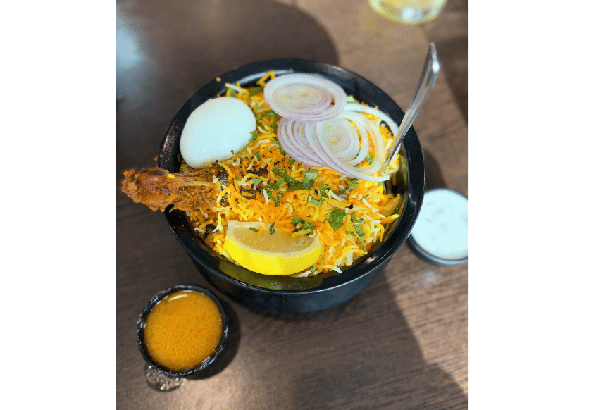 large black bowl filed with colorful yellow & orange basmati rice, topped with a lemon slice, red onion slices, a whole egg and green herbs.  dipping sauces on the side.  