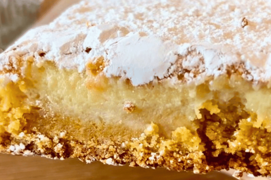 A zoomed in picture of a piece of deep dish gooey butter cake from Companion Cafe & Bakery.  brown & dark yellow crumbly cake on the bottom topped with a light yellow gooey layer.  Topped with a heavy layer of powdered sugar