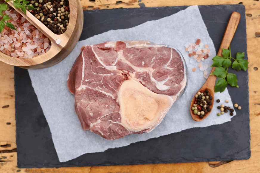 raw cut of beef, a cross cut shank, shown with bone-in and fat marbling.  course salt, herbs and whole peppercorns decorate the image with a spoonful of peppercorns framing the image.  You can get this grass-fed meat delivered from The Log Cabin Ranch