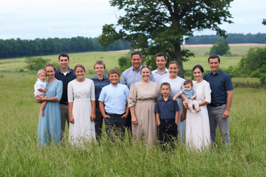 A large, 14 person, smiling family photo courtesy of The Log Cabin Ranch. The family is posing in the middle of a green pasture dressed in coordinating blues and neutrals 