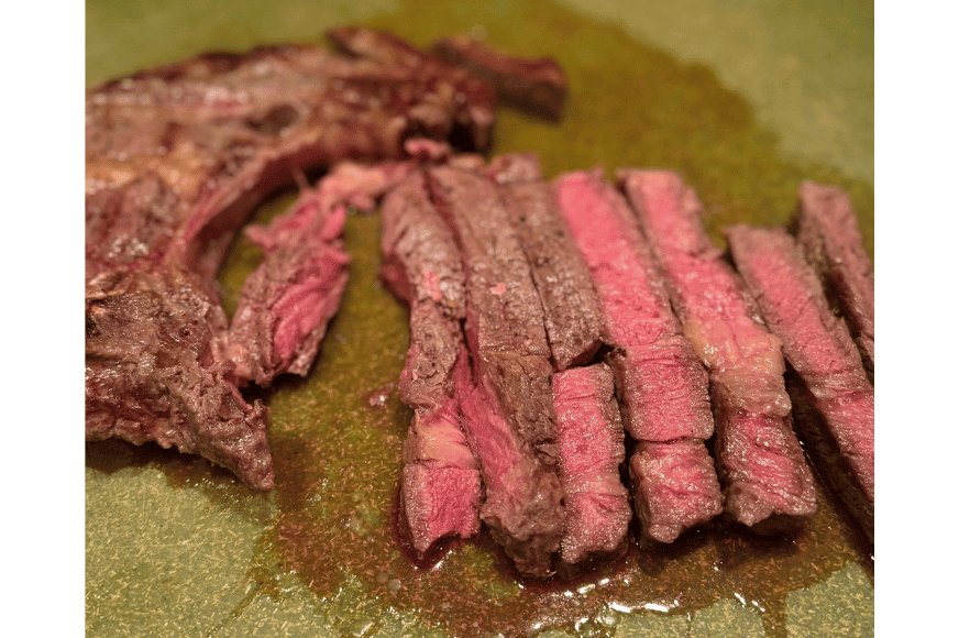 freshly sliced grass-fed meat that was delivered by The Log Cabin Ranch.  A bone-in ribeye steak on a green cutting board with juices running.  the steak is cooked medium temperature with pink inside.