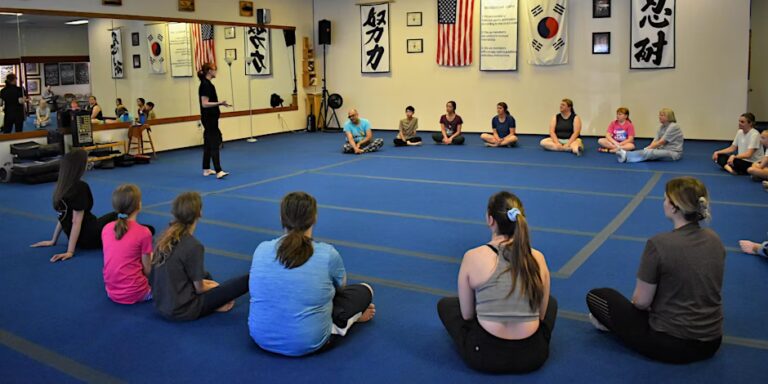 A group of women seated in a circle on a blue carpet in a martial arts studio