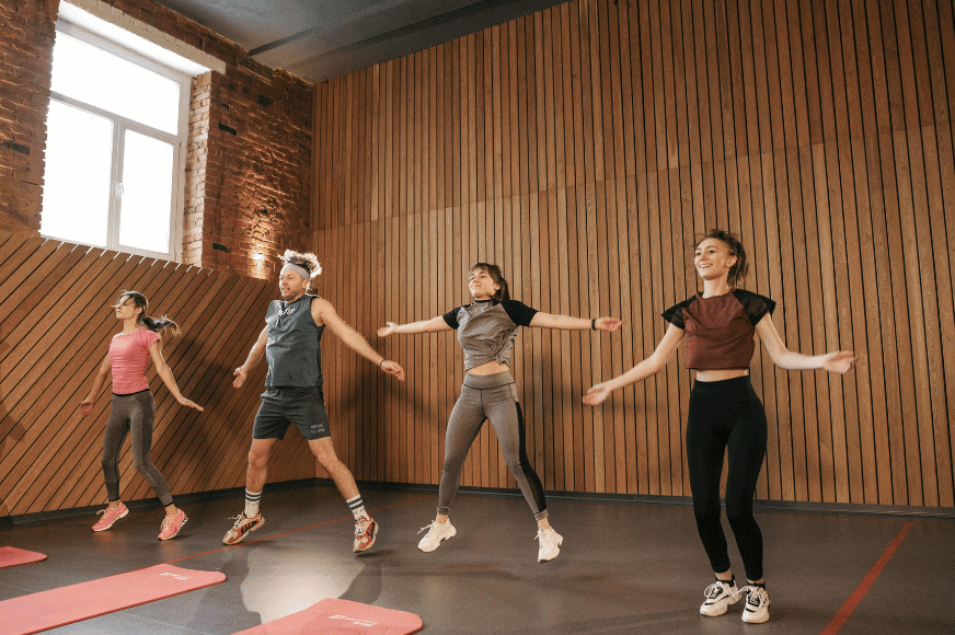 4 people doing a jumping jack in a group fitness studio with wooden back wall and concrete floors with red yoga mats laid out