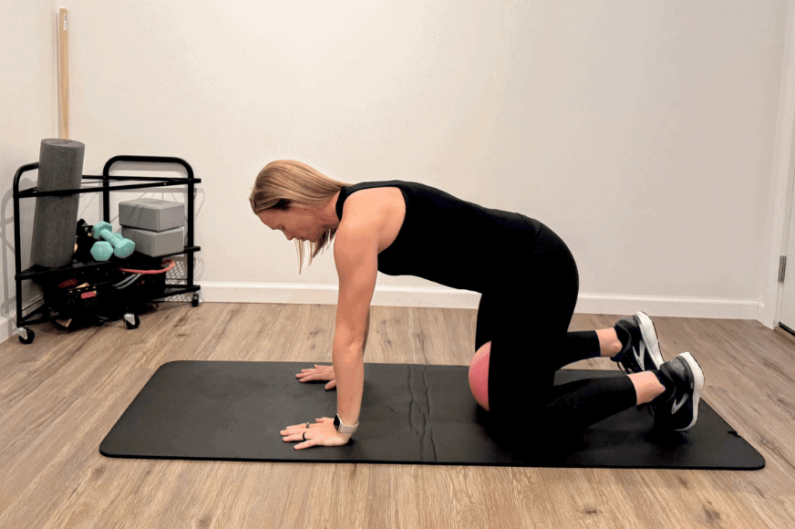 a blonde women wearing all black in table top position on a black yoga mat squeezes a red ball between her knees.