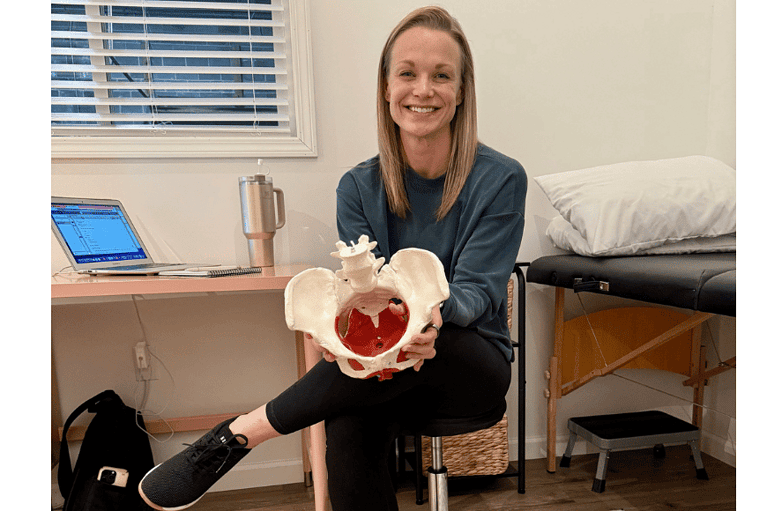 dr. Jamie Kocher, a blonde middle aged pelvic floor rehab specilist sits & smiles in her private office while holding the model of a women's pelvis including bones and muscles