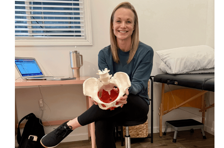 dr. Jamie Kocher, a blonde middle aged pelvic floor rehab specilist sits & smiles in her private office while holding the model of a women's pelvis including bones and muscles