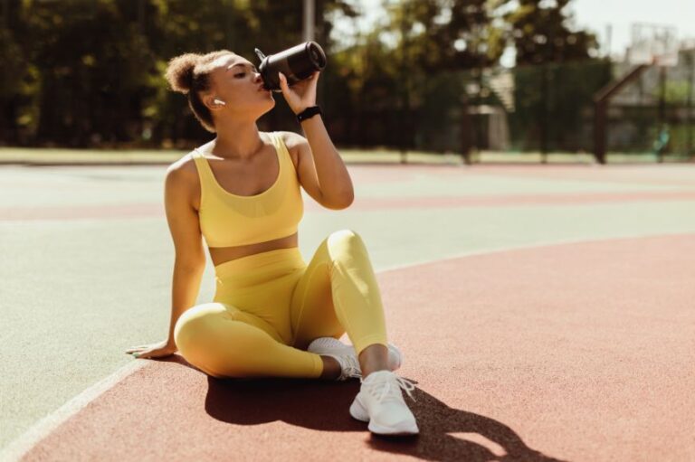women in yellow matching pants and sports bra consumes a coffee while resting on a track after a workout