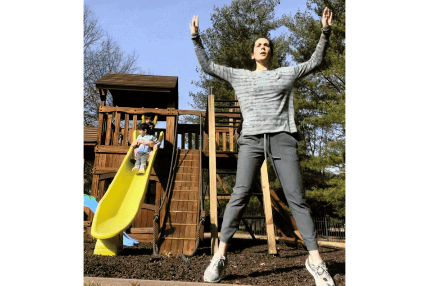 women doing a jumping jack while her child slides down the yellow slide behind her on the playground