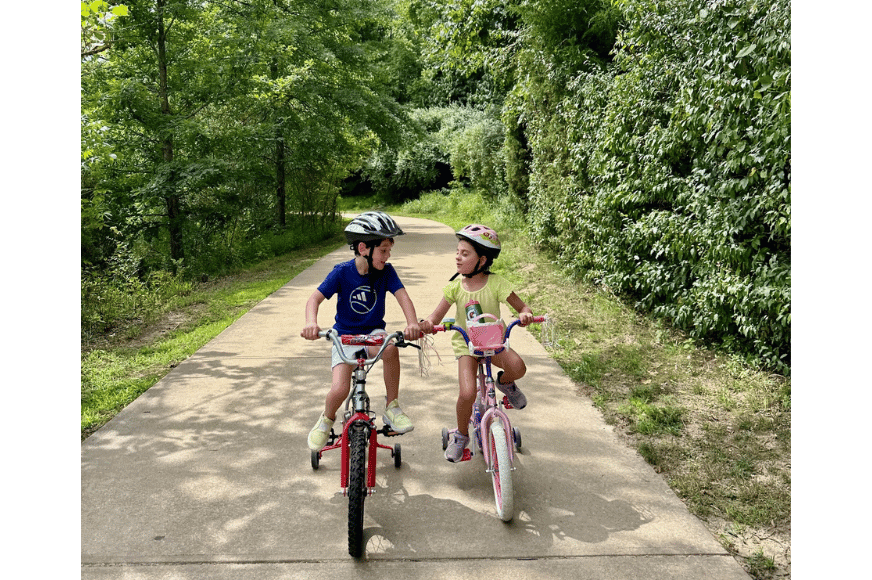 2 kids wearing helmuts biking along a paved path surrounded by green nature