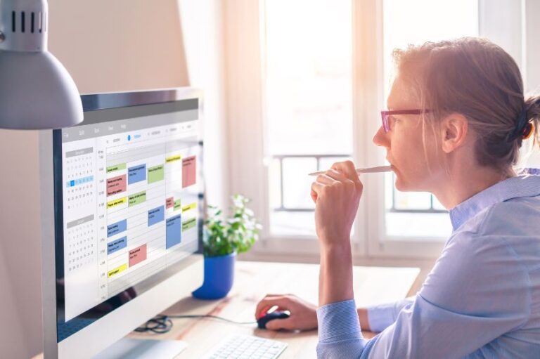 woman sitting at her desk looking at her busy online calendar at her desktop computer. she is holding a pen near her face. green plant in the background