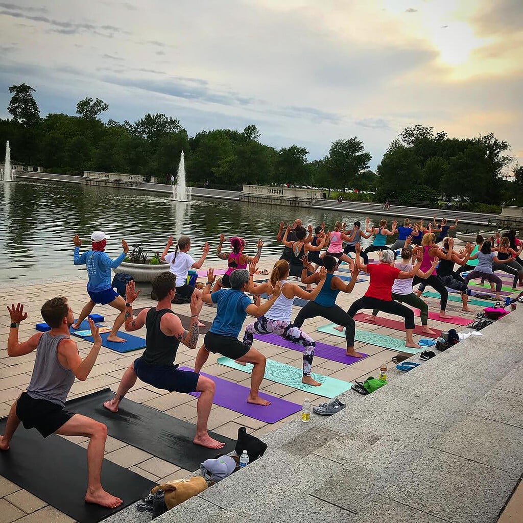 large group doing goddess pose at outdoor yoga by a lake with a fountain