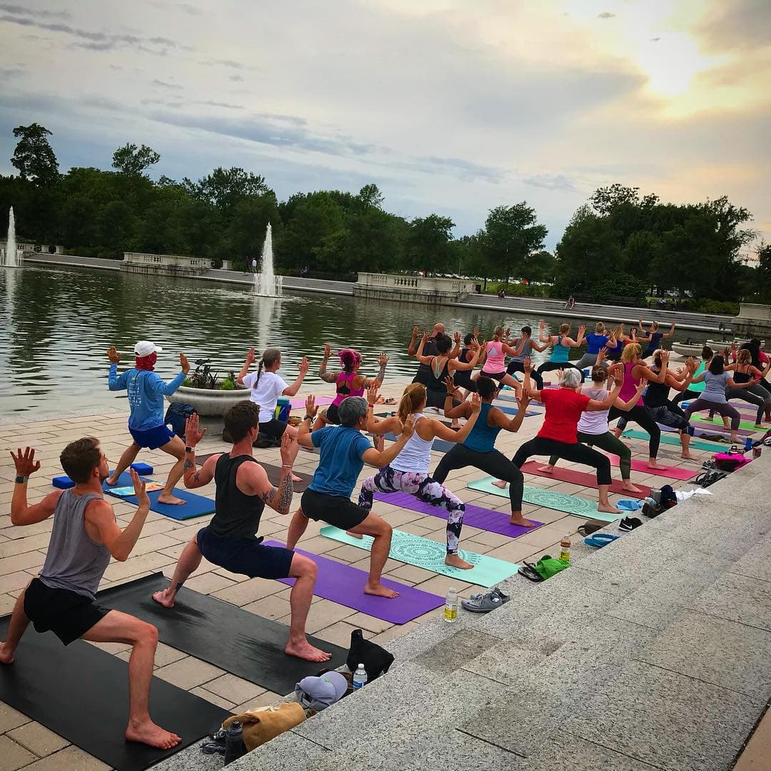 large group doing goddess pose at outdoor yoga by a lake with a fountain