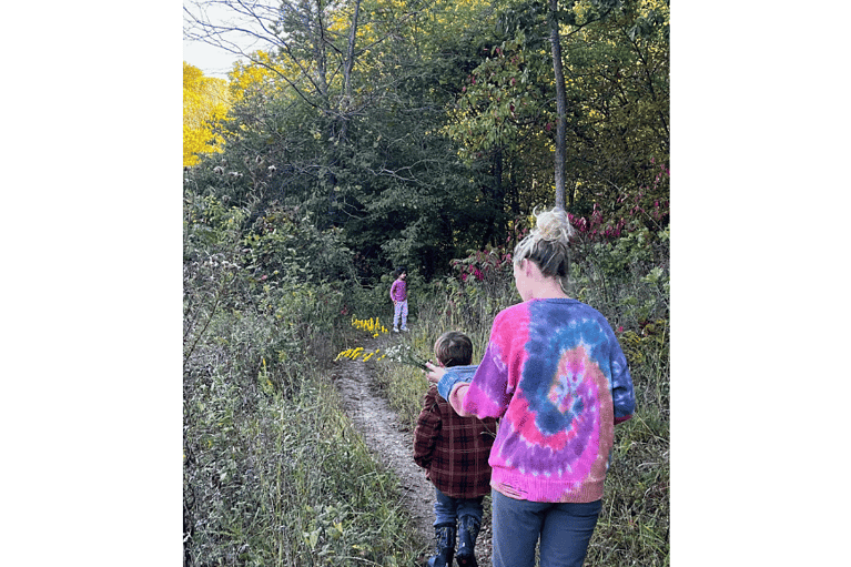 a mother and son hiking away along dirt path in grassy area with woods. another child is off in the distance