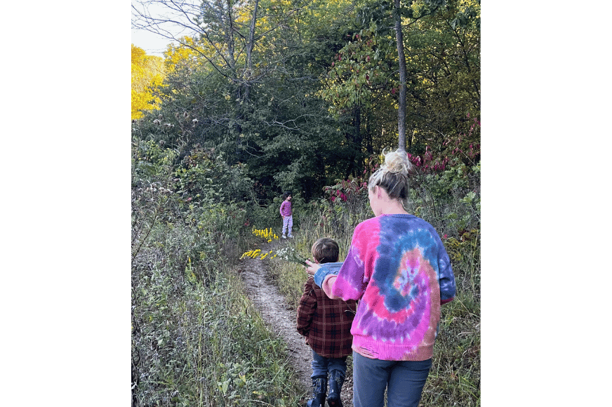 a mother and son hiking away along dirt path in grassy area with woods.  another child is off in the distance