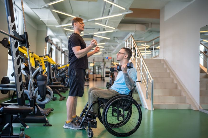 man in wheelchair lifting weights with guidance from his personal trainer at the gym