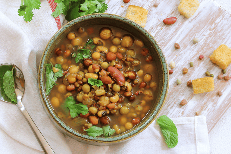 a bowl filled with bean soup - lentils, chickpeas and pinto beans in a broth shown with fresh cilantro and croutons on the side with a silver spoon.