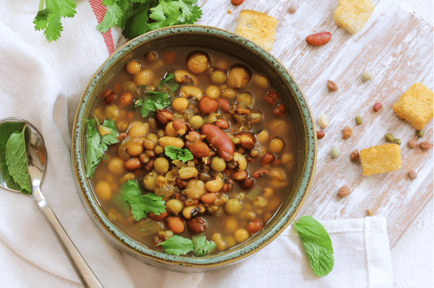 a bowl filled with bean soup - lentils, chickpeas and pinto beans in a broth shown with fresh cilantro and croutons on the side with a silver spoon.